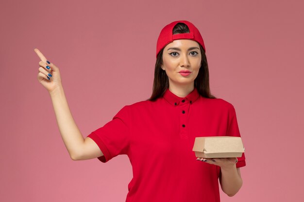 Vue de face jeune femme courrier en uniforme rouge et cape avec peu de colis de livraison de nourriture sur ses mains sur le mur rose