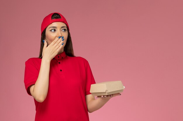 Vue de face jeune femme courrier en uniforme rouge et cape avec peu de colis de livraison de nourriture sur ses mains sur le mur rose