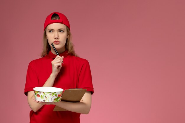 Vue de face jeune femme courrier en uniforme rouge et cape tenant le bol de livraison et le bloc-notes des notes d'écriture sur le mur rose