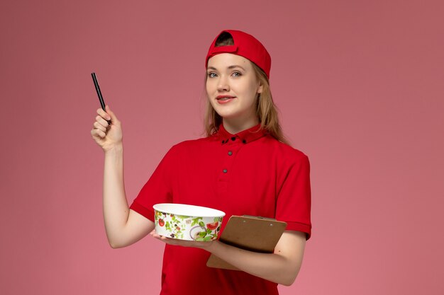 Vue de face jeune femme courrier en uniforme rouge et cape tenant le bol de livraison et le bloc-notes avec stylo mur rose