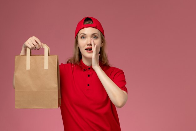 Vue de face jeune femme courrier en uniforme rouge et cape tenant le paquet alimentaire de livraison appelant sur le mur rose