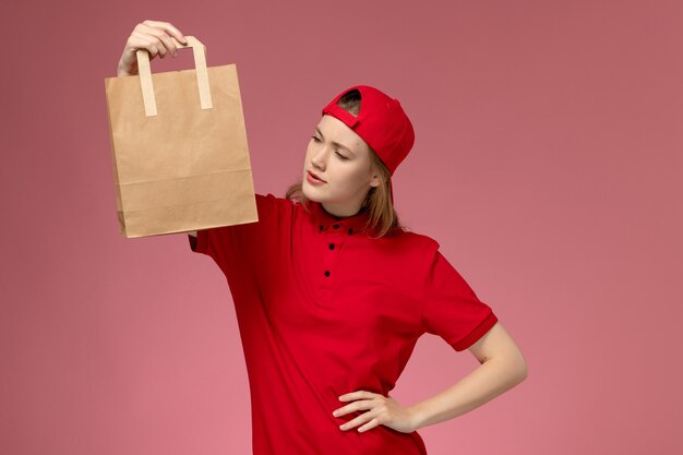 Vue de face jeune femme courrier en uniforme rouge et cape tenant le paquet de nourriture de livraison sur le mur rose