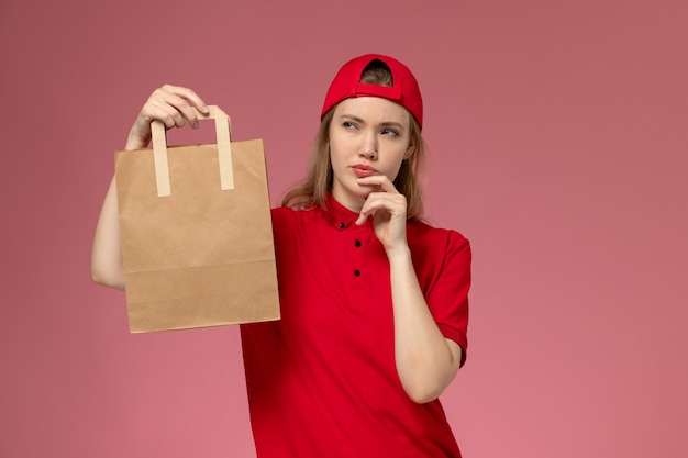 Vue de face jeune femme courrier en uniforme rouge et cape tenant le paquet de nourriture de livraison et la pensée sur le mur rose