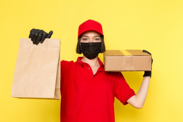 Une vue de face jeune femme courrier en uniforme rouge gants noirs masque noir et bonnet rouge tenant la boîte et le paquet de nourriture