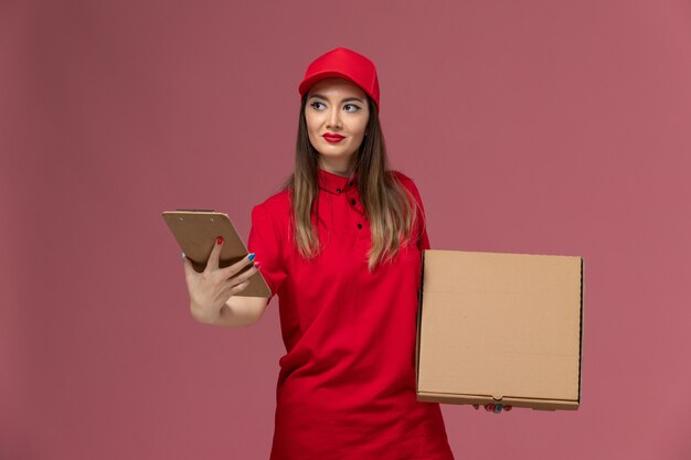 Vue de face jeune femme courrier en uniforme rouge tenant la boîte de nourriture de livraison avec bloc-notes sur fond rose clair service de livraison entreprise uniforme