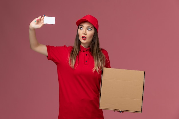 Vue de face jeune femme courrier en uniforme rouge tenant la boîte de nourriture de livraison et carte blanche sur le travail de l'entreprise uniforme de service de livraison de fond rose