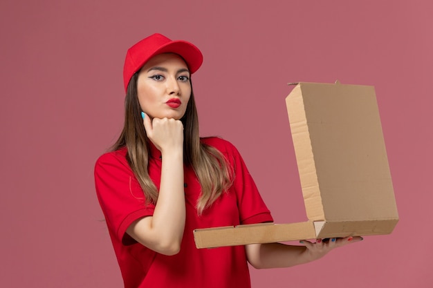 Vue de face jeune femme courrier en uniforme rouge tenant la boîte de nourriture de livraison sur fond rose entreprise de travail uniforme de livraison de services