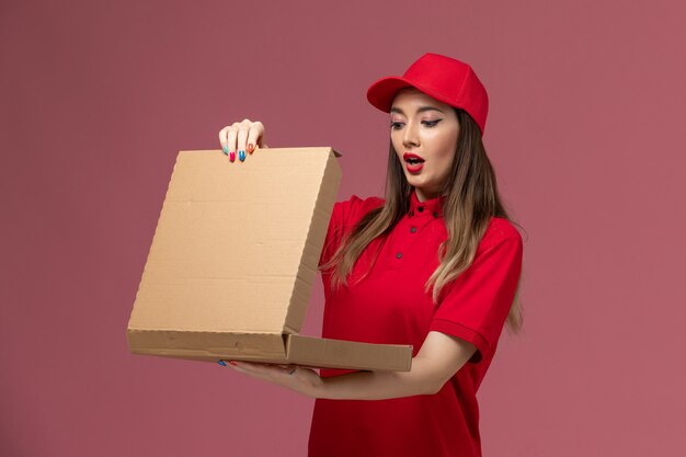 Vue de face jeune femme courrier en uniforme rouge tenant la boîte de nourriture de livraison de l'ouvrir sur fond rose service de livraison de l'entreprise uniforme de travail