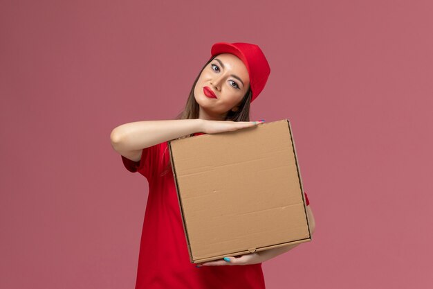 Vue de face jeune femme courrier en uniforme rouge tenant la boîte de nourriture de livraison sur le sol rose entreprise uniforme de livraison de services
