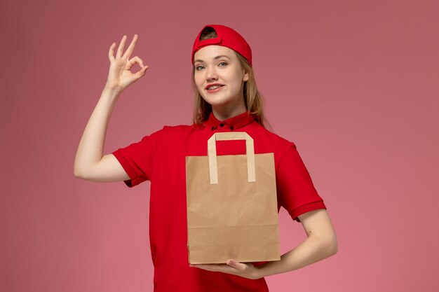 Vue de face jeune femme courrier en uniforme rouge tenant le paquet alimentaire de papier de livraison sur le mur rose clair