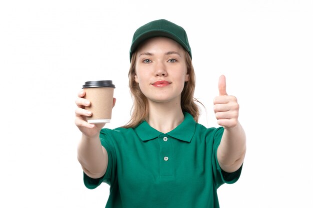 Une vue de face jeune femme courrier en uniforme vert tenant une tasse de café souriant et montrant comme signe sur blanc