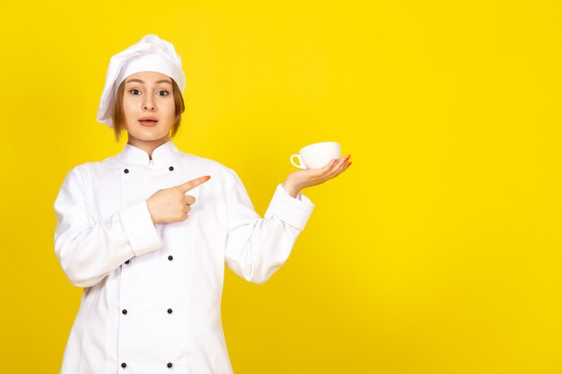 Une vue de face jeune femme cuisinier en costume de cuisinier blanc et bonnet blanc boire tenant une tasse de café sur le jaune