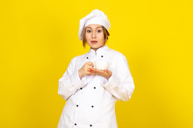 Une vue de face jeune femme cuisinier en costume de cuisinier blanc et bonnet blanc boire tenant une tasse de café sur le jaune