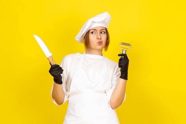 Une vue de face jeune femme cuisinier en costume de cuisinier blanc et bonnet blanc en gants noirs tenant un couteau et un nettoyant pour légumes sur le jaune