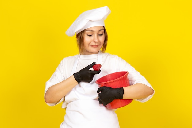 Une vue de face jeune femme cuisinier en costume de cuisinier blanc et bonnet blanc en gants noirs tenant panier rouge souriant sur le jaune