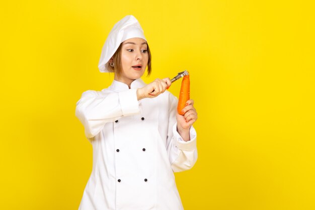Une vue de face jeune femme cuisinier en costume de cuisinier blanc et bonnet blanc tenant et nettoyage carotte orange sur le jaune