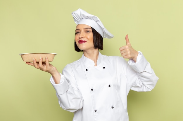 Une vue de face jeune femme cuisinier en costume de cuisinier blanc et cap holding bol avec sourire sur le mur vert