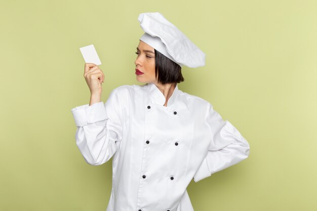 Une vue de face jeune femme cuisinier en costume de cuisinier blanc et cap holding carte blanche sur le mur vert dame travail couleur cuisine alimentaire