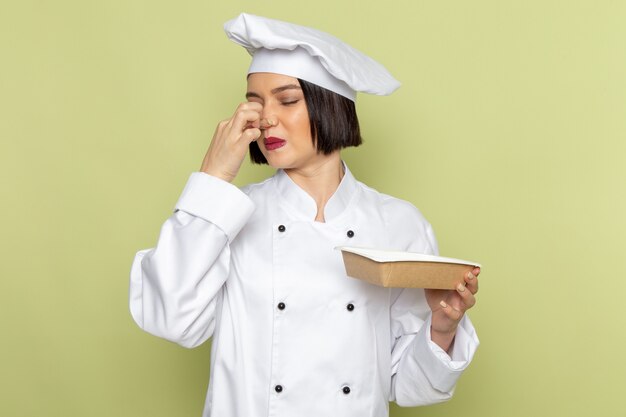 Une vue de face jeune femme cuisinier en costume de cuisinier blanc et cap holding package fermant son nez sur le mur vert dame travail couleur cuisine alimentaire