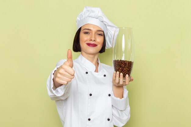 Une vue de face jeune femme cuisinier en costume de cuisinier blanc et cap holding pot avec des graines de café sur le mur vert dame travail couleur alimentaire