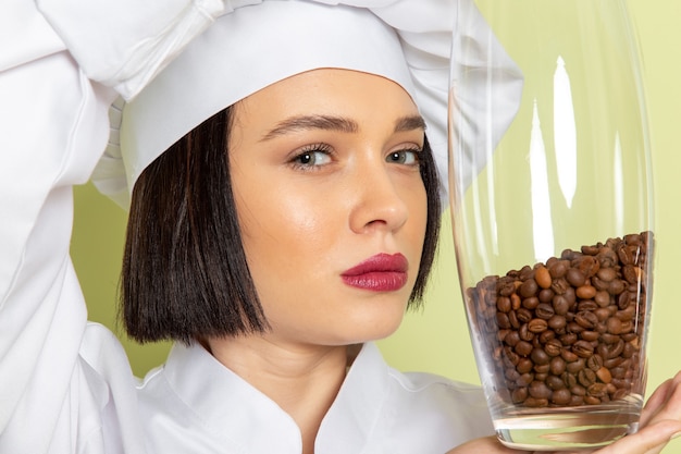 Photo gratuite une vue de face jeune femme cuisinier en costume de cuisinier blanc et cap holding pot avec des graines de café sur le mur vert dame travail cuisine couleur