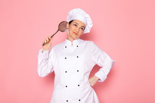 Une vue de face jeune femme cuisinier en costume de cuisinier blanc casquette blanche posant tenant une grosse cuillère en argent expression hésitante