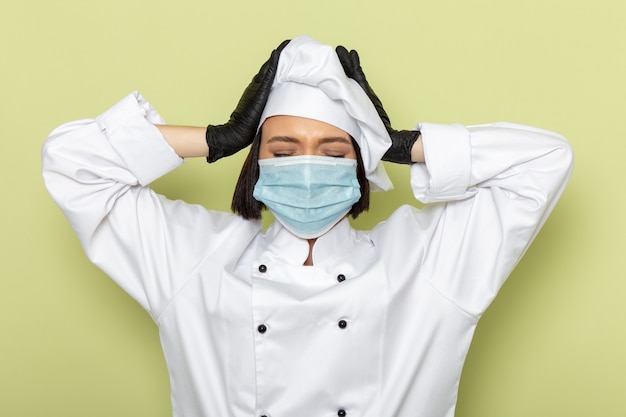 Une vue de face jeune femme cuisinier en costume de cuisinier blanc et casquette portant des gants et un masque stérile ayant de graves maux de tête sur le mur vert dame travail couleur alimentaire