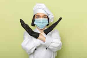 Photo gratuite une vue de face jeune femme cuisinier en costume de cuisinier blanc et casquette portant des gants et un masque stérile avec interdiction de poser sur le mur vert couleur alimentaire travail dame