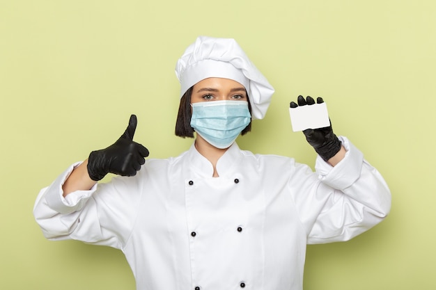 Une vue de face jeune femme cuisinier en costume de cuisinier blanc et casquette portant des gants et un masque stérile sur le mur vert dame travail couleur cuisine alimentaire