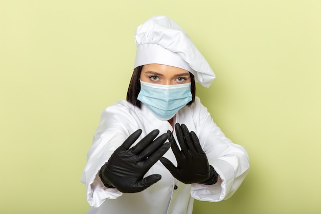 Photo gratuite une vue de face jeune femme cuisinier en costume de cuisinier blanc et casquette portant des gants et un masque stérile avec prudence pose sur le mur vert dame travail couleur alimentaire