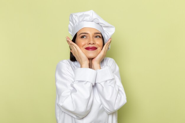 Une vue de face jeune femme cuisinier en costume de cuisinier blanc et chapeau posant avec une expression ravie sur le mur vert dame travail couleur cuisine alimentaire