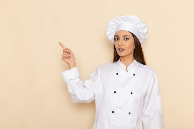 Vue de face de la jeune femme cuisinier en costume de cuisinier blanc levant son doigt sur un mur blanc clair