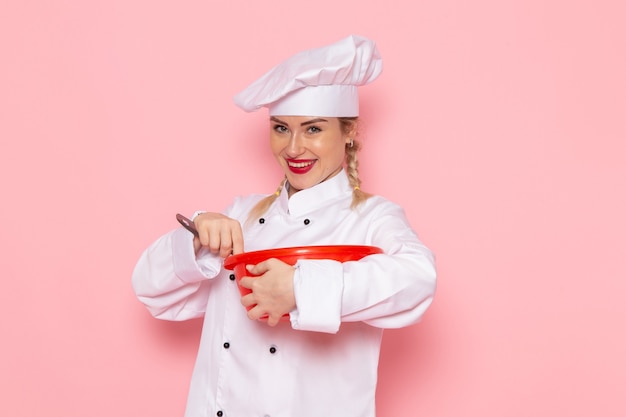 Photo gratuite vue de face jeune femme cuisinier en costume de cuisinier blanc mélangeant bol rouge sur l'espace rose cuisinier cuisine travail travail photo