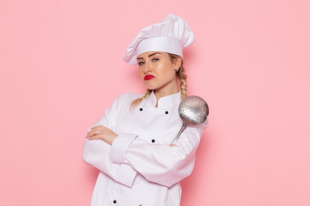 Vue de face jeune femme cuisinier en costume de cuisinier blanc posant avec une cuillère en argent sur l'espace rose cuisinier