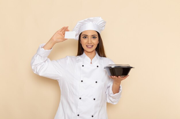 Vue de face de la jeune femme cuisinier en costume de cuisinier blanc tenant un bol noir et une carte sur un mur blanc clair