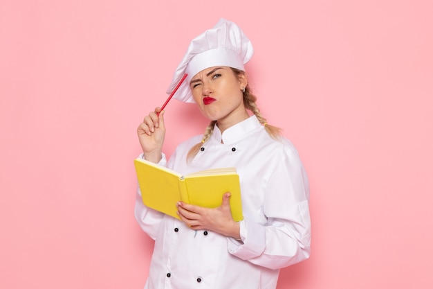 Vue de face jeune femme cuisinier en costume de cuisinier blanc tenant un cahier jaune et penser à l'espace rose cook