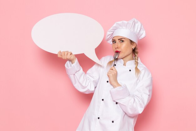 Vue de face jeune femme cuisinier en costume de cuisinier blanc tenant une pancarte blanche et cuillère en argent sur l'espace rose cook