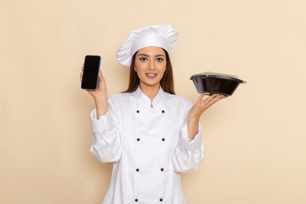 Vue de face de la jeune femme cuisinier en costume de cuisinier blanc tenant le téléphone et bol noir sur mur blanc