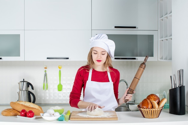 Vue de face jeune femme cuisinier pétrir la pâte sur une planche à découper tenant un rouleau à pâtisserie dans la cuisine