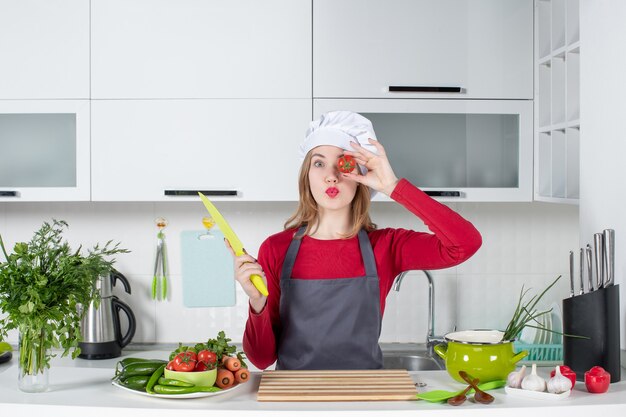 Vue de face jeune femme cuisinier en tablier tenant la tomate devant son œil