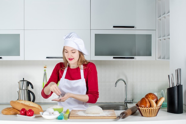 Vue de face jeune femme cuisinière en chapeau de cuisinier et tablier prenant de la farine dans un bol dans la cuisine