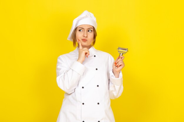 Une vue de face jeune femme cuisinière en costume de cuisinier blanc et capuchon blanc tenant un nettoyant pour légumes posant la pensée sur le jaune