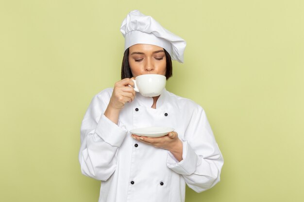 Une vue de face jeune femme cuisinière en costume de cuisinier blanc et capuchon de boire du café sur le mur vert