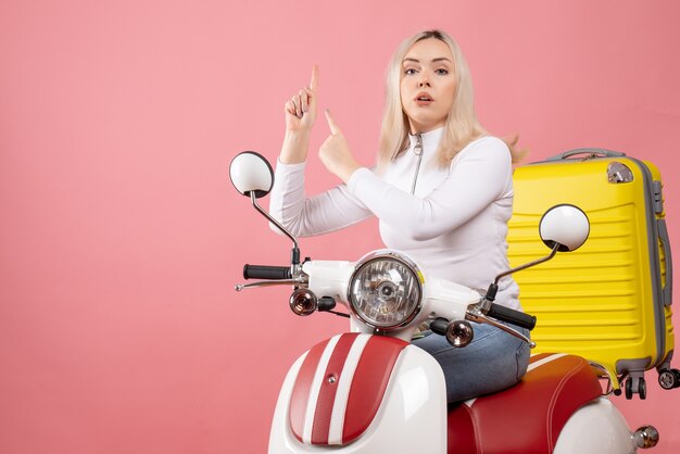 Vue de face jeune femme sur cyclomoteur avec valise jaune pointant les doigts vers le haut