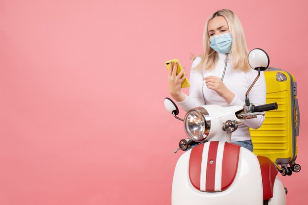 Vue de face jeune femme sur cyclomoteur avec valise jaune en regardant son téléphone