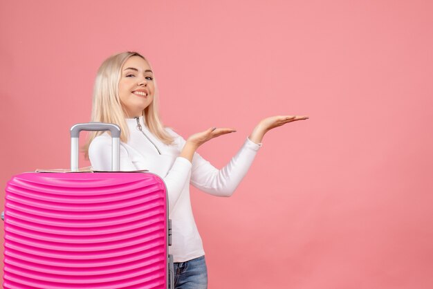 Vue de face jeune femme debout derrière une valise rose pointant vers la gauche