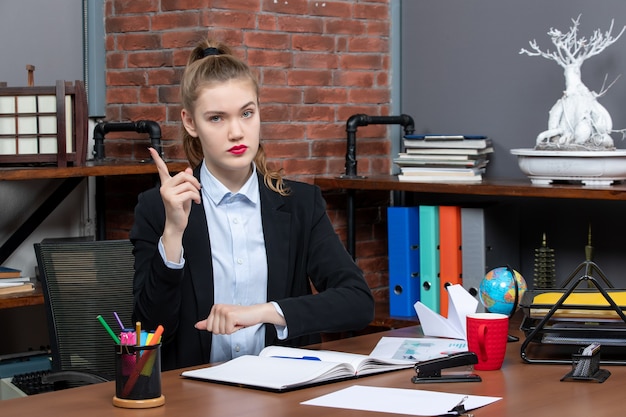 Vue de face d'une jeune femme déterminée assise à une table et pointant vers le haut dans le bureau