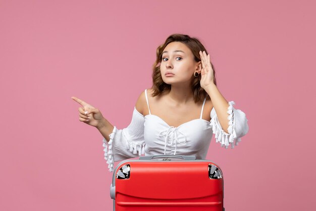 Vue de face d'une jeune femme écoutant attentivement avec un sac de vacances rouge sur un mur rose