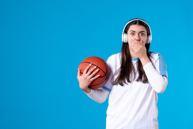 Vue de face jeune femme avec des écouteurs tenant le basket-ball sur le mur bleu
