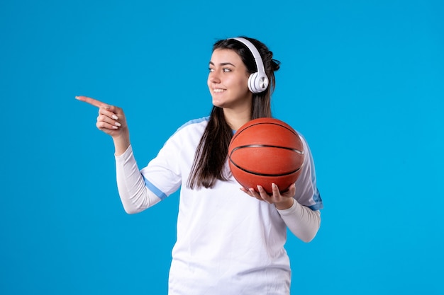 Vue de face jeune femme avec des écouteurs tenant le basket-ball sur le mur bleu